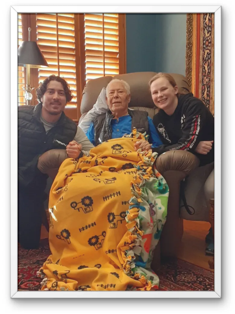 Family photo from Our Story: two family members kneeling beside an older man seated in an armchair under a blanket